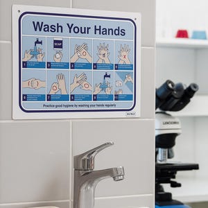 May include: A sign instructing hand washing is fixed to a tiled wall above a sink. The sign features blue and white text and diagrams. A chrome tap and a bar of soap are also visible in the image.