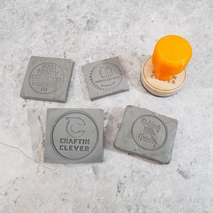 May include: Five gray square ceramic stamps with different designs. The stamps are on a gray surface. The designs include "Sam Henry Ceramics Melbourne AU", "Mitraceramics Handmade", "C Craftin Clever", "Baking With Trinity", and a stamp with a circle design.