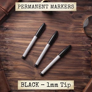 May include: Three permanent markers with light grey barrels and black caps, featuring a 1mm tip. The text PERMANENT MARKERS and BLACK - 1mm Tip is displayed on the image. These writing instruments are arranged diagonally on a dark wood surface.