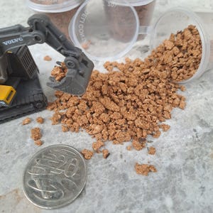 May include: A toy digger scoops up brown, granular material, next to a coin. The digger is black and yellow, with the Volvo logo visible. Several clear plastic containers with lids are in the background, some containing the same brown material.