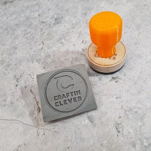 May include: A gray rubber stamp with the text "Craftin Clever" and a stylized letter "C" inside a circle. The stamp is next to an orange rubber stamp handle.