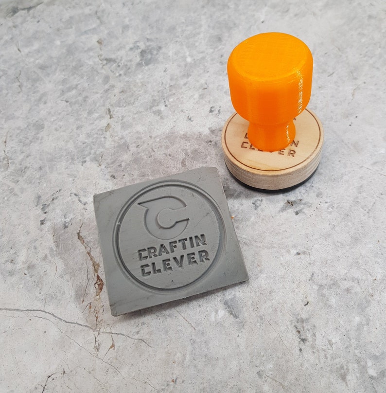 May include: A gray rubber stamp with the text "Craftin Clever" and a stylized letter "C" inside a circle. The stamp is next to an orange rubber stamp handle.