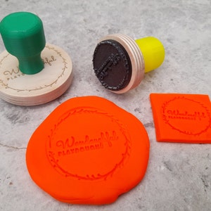 Puede incluir: Tres sellos de madera con el texto "Wonderful Playground" en un dise&ntilde;o circular. Los sellos son verde, negro y naranja. El sello naranja est&aacute; sobre una plastilina naranja brillante.