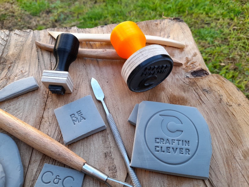 May include: A close-up of a wooden surface with various tools for crafting. There are two stamps, one with a black handle and the other with an orange handle. There are also two tools with wooden handles and a metal scraper. There are four pieces of gray clay with different designs stamped into them. The designs include "REMA", "C&C", and "CRAFTIN CLEVER".