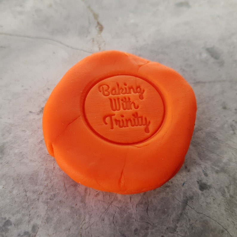 Puede incluir: Masilla naranja con una impresi&oacute;n redonda que dice "Baking With Trinity" en blanco.