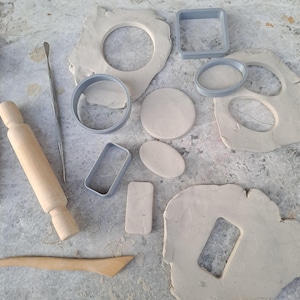 Può includere: Una collezione di strumenti per scolpire l'argilla e pezzi di argilla. Include un mattarello di legno, una spatola di metallo e vari tagliaforme in plastica grigia. L'argilla è beige chiaro, con forme ritagliate di cerchi, quadrati e ovali.