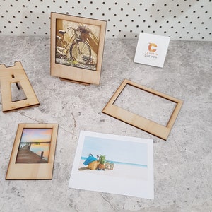Peut inclure: Un ensemble de quatre cadres photo en bois avec un fond blanc et une finition en bois clair. Les cadres sont en forme d'appareil photo instantan&eacute; Polaroid. Les cadres sont de tailles diff&eacute;rentes et sont dispos&eacute;s en rang&eacute;e sur une surface gris clair. Les cadres sont parfaits pour afficher des photos ou des &oelig;uvres d'art.