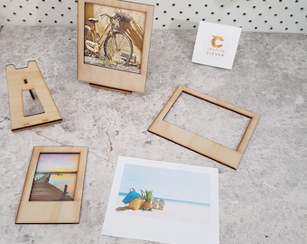 Puede incluir: Un conjunto de cuatro marcos de fotos de madera con fondo blanco y acabado en madera clara. Los marcos tienen la forma de una cámara instantánea Polaroid. Los marcos son de diferentes tamaños y están dispuestos en fila sobre una superficie gris claro. Los marcos son perfectos para mostrar fotos u obras de arte.