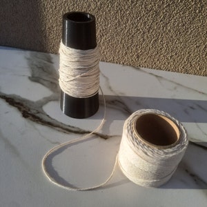 Könnte beinhalten: Zwei Spulen Naturfarbener Schnur. Eine Spule ist um einen schwarzen, zylindrischen Halter gewickelt. Die andere Spule ist eine Rolle Schnur. Die Schnur hat eine hellbeige Farbe und scheint aus Naturfasern zu bestehen.