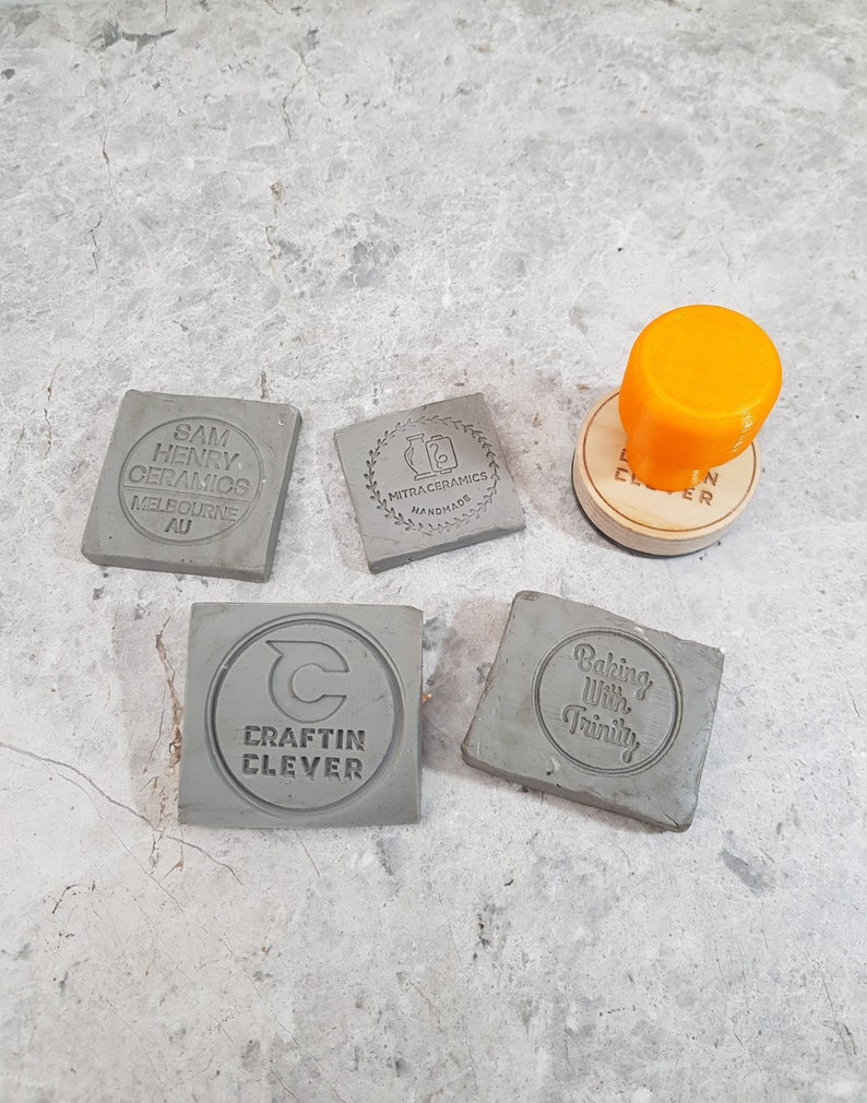 May include: Five gray square ceramic stamps with different designs. The stamps are on a gray surface. The designs include "Sam Henry Ceramics Melbourne AU", "Mitraceramics Handmade", "C Craftin Clever", "Baking With Trinity", and a stamp with a circle design.