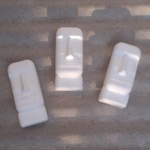 May include: Three white Moai statues, each with a rectangular shape and carved facial details, including a nose and base. They are positioned on a textured, grey surface, with shadows adding depth.