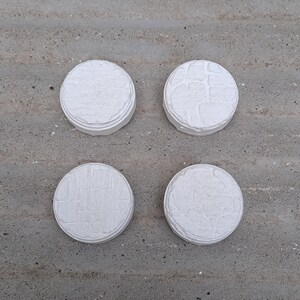 May include: Four white round ceramic tiles with different raised patterns. The tiles are arranged in a square pattern on a grey surface.