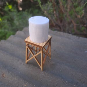 May include: A white plastic water tower sits atop a wooden stand with a simple X-shaped design. The tower is approximately 25 cm tall.