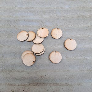 May include: A selection of unfinished, circular wooden tags with small pre-drilled holes. The tags are light brown and spread across a grey surface. These craft supplies are suitable for DIY projects, personalisation, or embellishments.