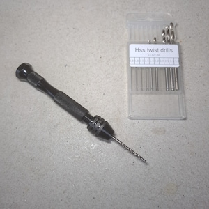 May include: A black drill bit holder with a drill bit inserted. The holder has a textured grip and a chuck for holding the drill bit. A clear plastic case with a label that reads "Hss twist drills size:mm" contains a set of drill bits.