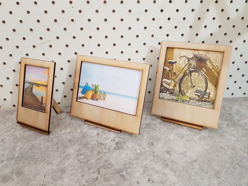 Peut inclure: Trois cadres photo en bois avec des images diff&eacute;rentes. Les cadres sont tous debout sur une surface grise. L'image dans le plus grand cadre est d'un v&eacute;lo avec un panier de fleurs. L'image dans le cadre du milieu est d'une sc&egrave;ne de plage avec un ananas et une serviette bleue. L'image dans le plus petit cadre est d'un coucher de soleil sur un lac avec une jet&eacute;e.