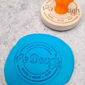 Puede incluir: Una masa de juego azul con un sello redondo de madera que dice "123 Dough The Original Shake Make Play" en un c&iacute;rculo.