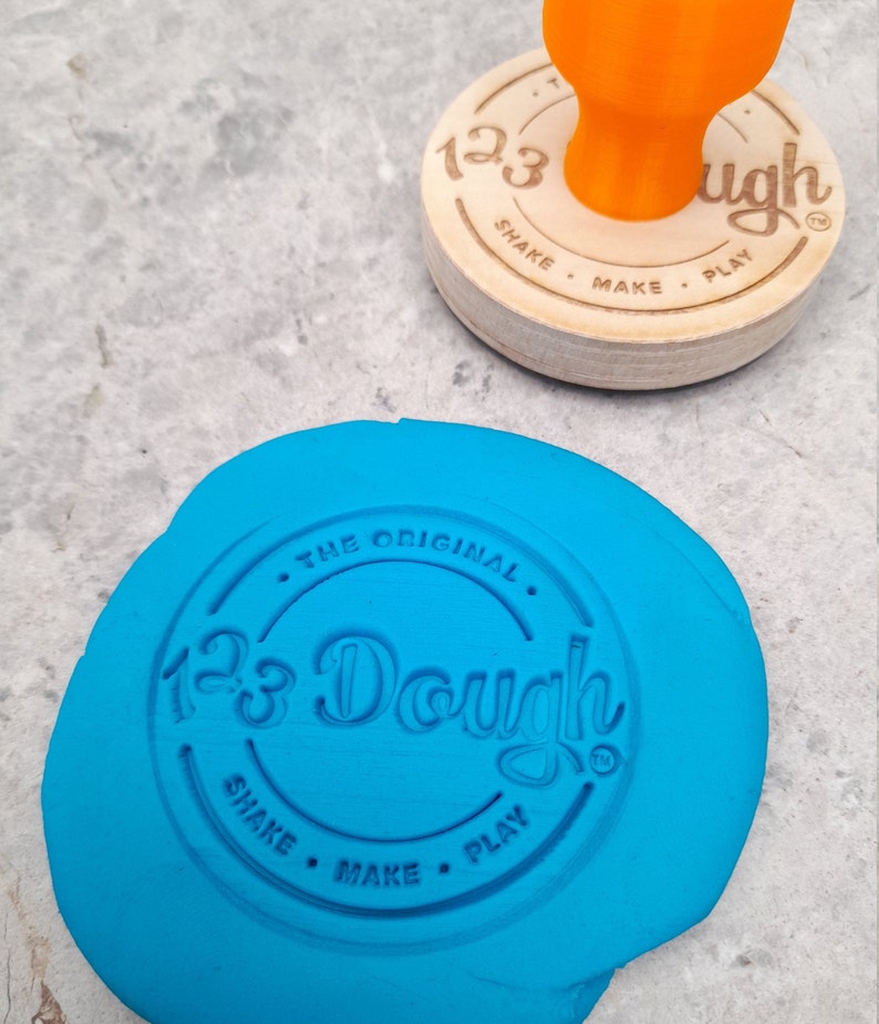 Puede incluir: Una masa de juego azul con un sello redondo de madera que dice "123 Dough The Original Shake Make Play" en un c&iacute;rculo.