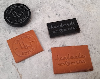 May include: An assortment of leather craft items, including a round black coaster with "LEATHER WORKS" text, and two rectangular tags with "handmade with ♡ by ALEXA" and "LEATHER WORKS" embossed. The items are in shades of brown and black.
