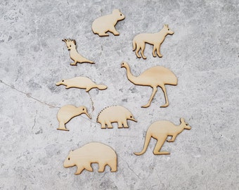 Puede incluir: Un conjunto de nueve recortes de animales de madera, que incluyen un koala, un dingo, un emú, un ornitorrinco, un kiwi, un equidna, un canguro, un wombat y un cacatúa. Los recortes están hechos de madera marrón claro y están dispuestos sobre un fondo gris.