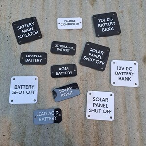 May include: A collection of black and white rectangular and oval labels with text. The labels include phrases like "BATTERY MAIN ISOLATOR", "CHARGE CONTROLLER", and "SOLAR PANEL SHUT OFF".