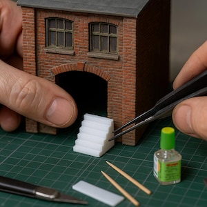 Peut inclure: Maquette d'un bâtiment en briques avec une entrée en arc et deux fenêtres. Un petit escalier blanc est assemblé avec une pince à épiler. D'autres outils incluent une bouteille de colle, des cure-dents et un petit morceau rectangulaire.