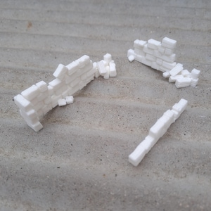 May include: White miniature brick wall sections, some intact and others broken, scattered on a textured grey surface. The pieces are arranged to resemble a partially demolished structure, ideal for model building or crafts.