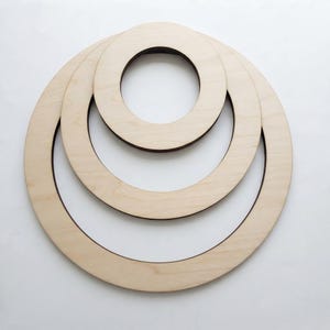 May include: Three nested circular wooden frames made of light-coloured plywood. The rings are arranged concentrically, with the smallest ring at the top, a medium ring in the centre, and the largest ring forming the outer base of the structure.