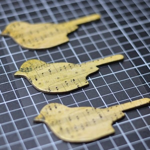 May include: Three yellow paper bird cutouts with musical notes printed on them. The birds are arranged in a row on a black and white grid background.