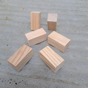 Può includere: Sei blocchi di legno non rifiniti di varie dimensioni e forme sono disposti su una superficie grigia. I blocchi sono prismi rettangolari con tagli angolati su un lato, che mostrano la venatura e il colore naturale del legno.