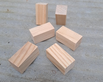May include: Six unfinished wooden blocks of varying sizes and shapes are arranged on a gray surface. The blocks are rectangular prisms with angled cuts on one side, showcasing the natural wood grain and color.