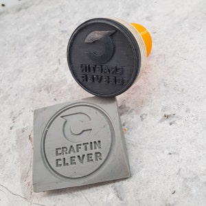 May include: A black rubber stamp with the text "CRAFTIN CLEVER" in reverse lettering and a stylized "C" logo. The stamp is mounted on a yellow plastic handle. A gray rubber stamp with the same text and logo is also shown.