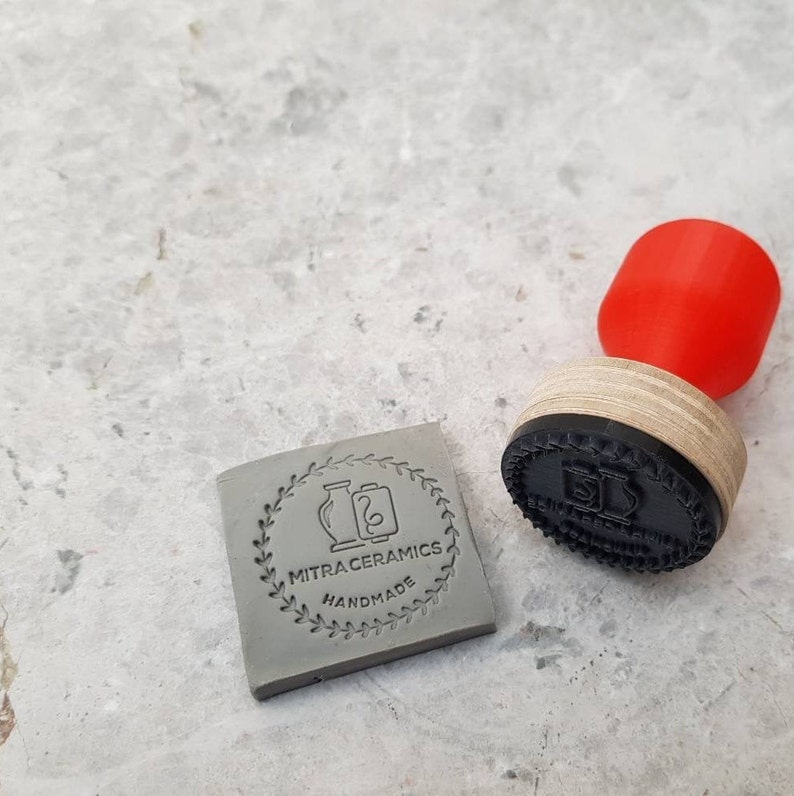 May include: A gray clay stamp with a circular design featuring two vases and the text "MITRACERAMICS HANDMADE". The stamp is next to a red rubber stamp with a black wooden handle.