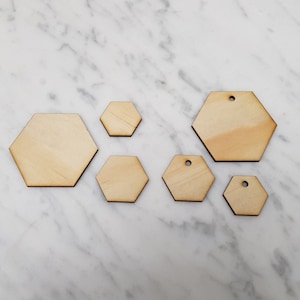 May include: Six unfinished wooden hexagon shapes of varying sizes. The hexagons have a natural wood color and are arranged on a white marble surface.