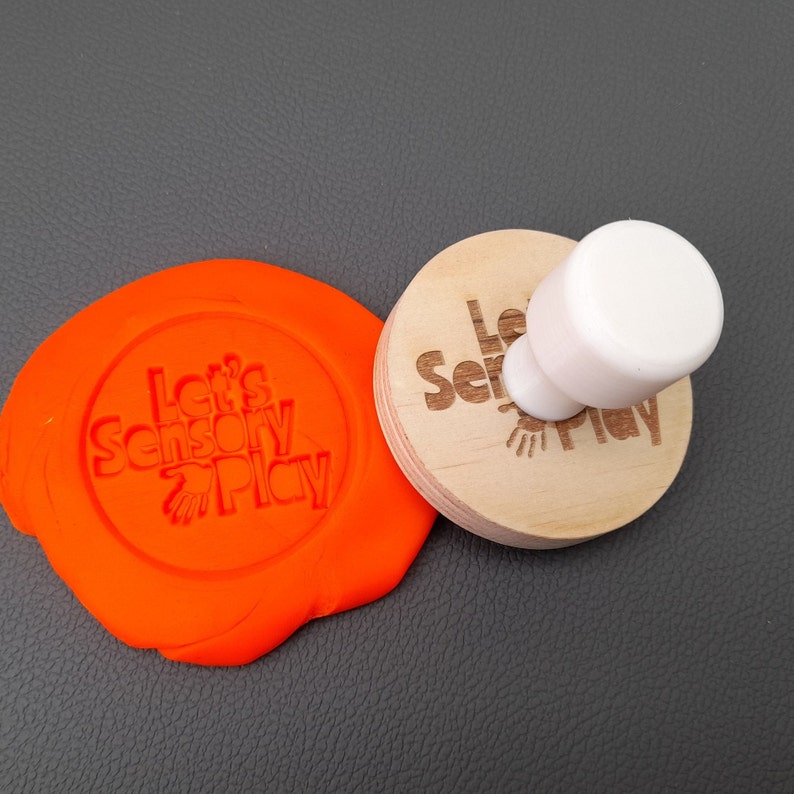 Puede incluir: Un sello de madera con el texto "Let's Sensory Play" y un dise&ntilde;o de huella de mano. El sello se est&aacute; utilizando para crear una impresi&oacute;n en una masa de juego naranja brillante.