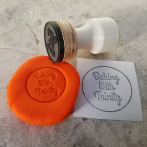 Puede incluir: Un sello de goma con el texto "Baking With Trinity" sobre un fondo blanco. El sello est&aacute; al lado de una masa de juego naranja con el mismo texto impreso.