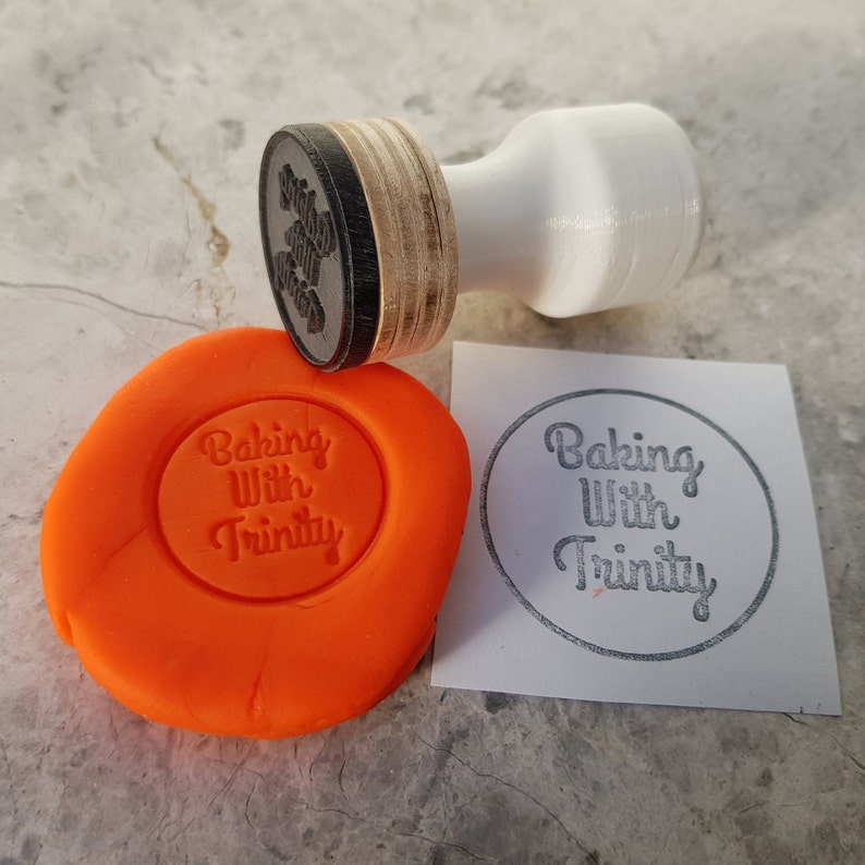 Puede incluir: Un sello de goma con el texto "Baking With Trinity" sobre un fondo blanco. El sello est&aacute; al lado de una masa de juego naranja con el mismo texto impreso.
