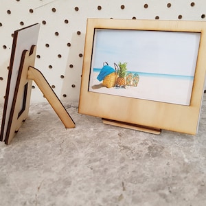 Peut inclure: Deux cadres photo en bois avec un support. Le cadre de droite contient une photo d'une sc&egrave;ne de plage avec un ananas, une serviette de plage et des tongs.