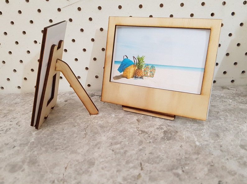 Peut inclure: Deux cadres photo en bois avec un support. Le cadre de droite contient une photo d'une sc&egrave;ne de plage avec un ananas, une serviette de plage et des tongs.