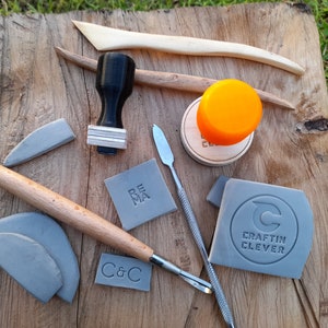 May include: A collection of pottery tools and stamps, including a rolling pin, a wooden stamp with the word "REMA" on it, a gray stamp with the text "CRAFTIN CLEVER" and a circle with a "C" inside, and a gray stamp with the text "C&C".