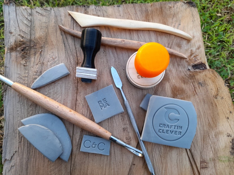 May include: A collection of pottery tools and stamps, including a rolling pin, a wooden stamp with the word "REMA" on it, a gray stamp with the text "CRAFTIN CLEVER" and a circle with a "C" inside, and a gray stamp with the text "C&C".