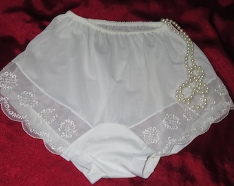 May include: White nylon panties with a scalloped lace trim and embroidered floral designs. The panties are on a red background with a pearl necklace.