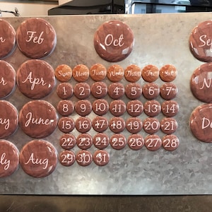 May include: A magnetic calendar with brown wooden-look month and date markers. The months are in a cursive font. The calendar is on a silver metal background. The dates are in a simple font.