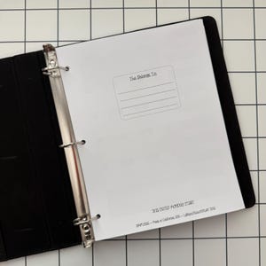Puede incluir: Una carpeta negra de tres anillas que contiene una página blanca con un campo "This Belongs To:", cuatro líneas en blanco y el texto "2026 DATED MONDAY START" seguido de "SIMPLIFY - Made in California, USA -- LilyPandaStickerStuff 2026" abajo.