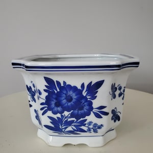 Könnte beinhalten: Weißer Keramik-Pflanztopf mit blauem Blumendesign. Der achteckige Pflanztopf hat ein blaues Band am Rand und blaue Blumen- und Schmetterlingsmotive. Der Pflanztopf hat einen Fuß und ist für den Innen- und Außenbereich geeignet.