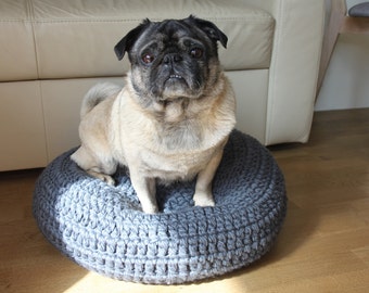 May include: A pug dog sitting on a gray crocheted dog bed. The dog is looking at the camera.