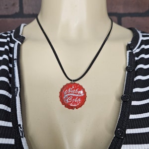 May include: A black cord necklace with a red bottle cap pendant. The bottle cap has the words "Nuka Cola" printed on it in white.