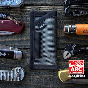 Può includere: Una custodia in nylon grigia con un cinturino nero e il testo "ARC Co." stampato su di essa. La custodia è progettata per contenere un multi-tool o un altro piccolo coltellino da tasca. La custodia è su una superficie di legno con altri strumenti e coltelli.
