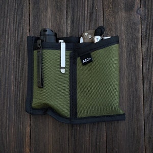 May include: A green zippered pouch with black trim and a black label that says "ARC Co.". The pouch is filled with a black flashlight, a silver pen, and a silver pocket knife.