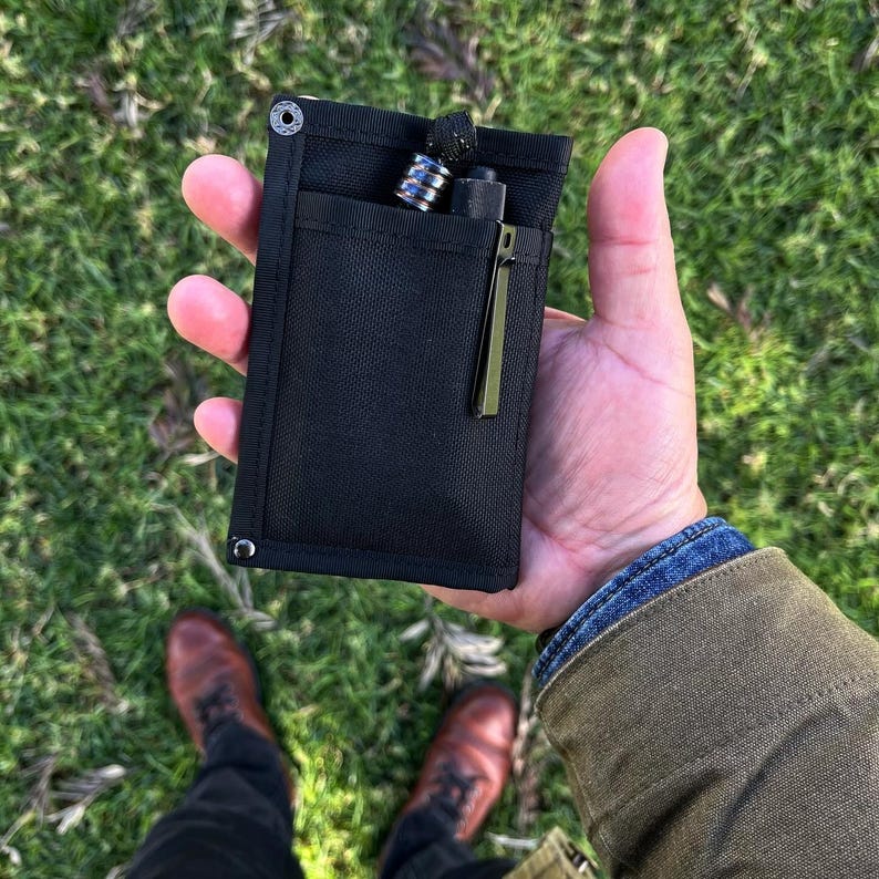 May include: A black fabric pouch with a zippered pocket and two metal snaps. The pouch is holding a small silver pen and a black pen.